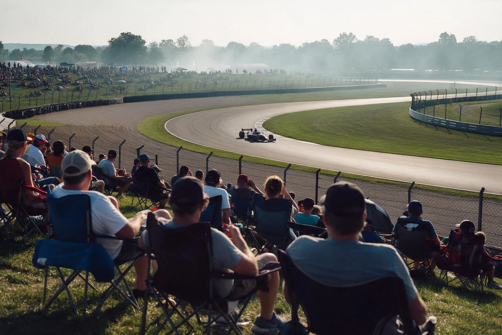 Conceptual illustration of spectators on a grassy general-admission bank watching a single-seater pass through a corner below them