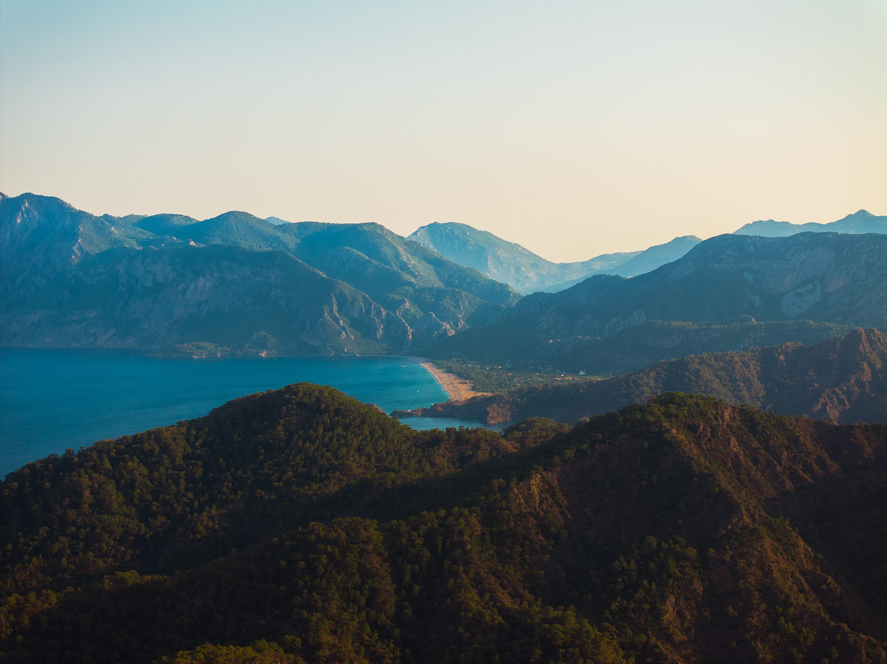 Forested hills and a bay on the Kemer coast at sunset, with the mountains beyond